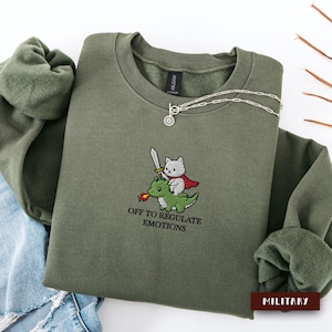 May include: An olive green crewneck sweatshirt featuring an embroidered graphic of a white cat wearing a red cape and holding a sword while riding a green dragon breathing fire. Below the graphic is the text "OFF TO REGULATE EMOTIONS".