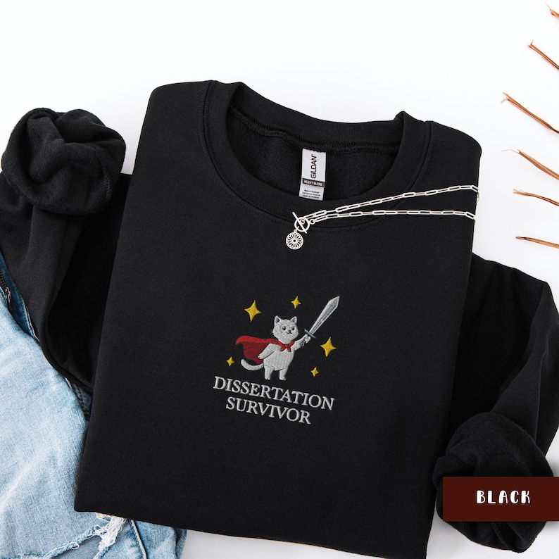 May include: Black sweatshirt with a crew neck. The sweatshirt features an embroidered design of a cat wearing a cape and holding a sword, with the text "Dissertation Survivor" below. A silver necklace is draped across the sweatshirt.