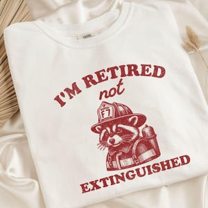 May include: White t-shirt with a red graphic of a raccoon wearing a firefighter helmet and gear. The text reads "I'M RETIRED not EXTINGUISHED" in a vintage style font.