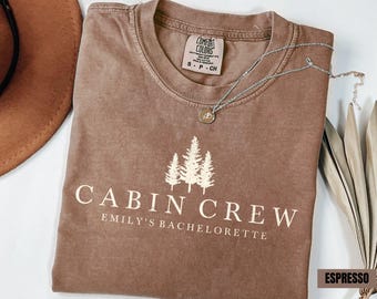 May include: Espresso-colored t-shirt with the text "CABIN CREW" and "Emily's Bachelorette" in white. The shirt features a graphic of three pine trees. A silver necklace with a round pendant rests on the shirt.