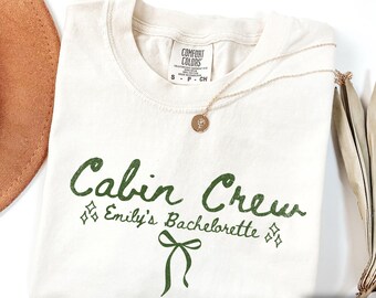 May include: Ivory-colored t-shirt with the text "Cabin Crew Emily's Bachelorette" in green. The shirt has a crew neck and short sleeves. A gold necklace with a pendant rests on the shirt. A brown hat is visible in the upper left corner.