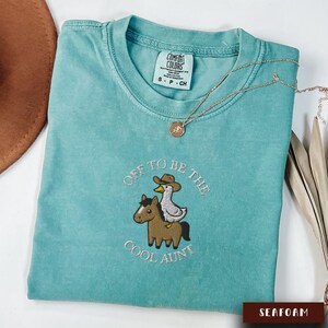 May include: Seafoam green Comfort Colors t-shirt with the embroidered phrase "Off to be the Cool Aunt." The design features a horse and a duck wearing a cowboy hat. A gold necklace with a pendant is draped on the shirt.
