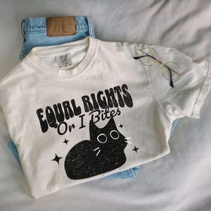 May include: A cream-colored t-shirt with the text "EQUAL RIGHTS Or I Bites" and a black cat graphic. The shirt is paired with blue jeans and round-framed glasses.