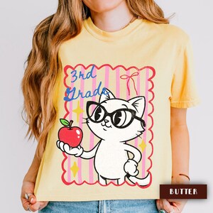 Puede incluir: Camiseta amarilla con un gato de dibujos animados con gafas y una manzana roja. El diseño incluye el texto "3rd Grade" en azul y rosa, dentro de un marco rosa y rojo con rayas verticales y estrellas.