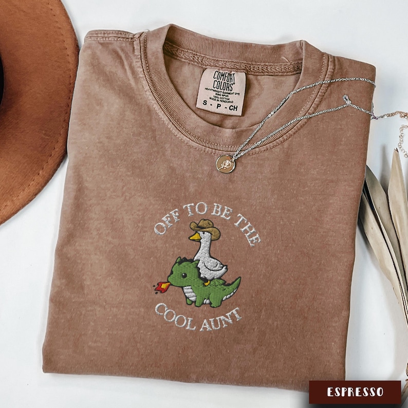 May include: Espresso-colored t-shirt with the phrase "Off to be the Cool Aunt" embroidered on the front. The design features a duck wearing a cowboy hat riding a green dinosaur. A silver necklace is draped across the shirt.