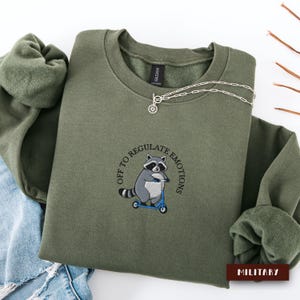 May include: An olive green crewneck sweatshirt featuring an embroidered graphic of a raccoon riding a blue scooter. The text "OFF TO REGULATE EMOTIONS" is arched above the raccoon. A silver-toned chain necklace rests on the neckline.