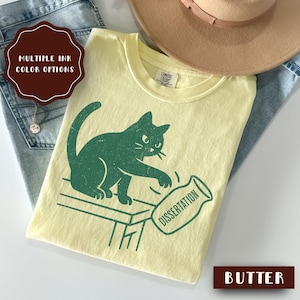 May include: Pale yellow t-shirt featuring a green cat knocking over a jar with the word "Dissertation." The shirt has a crew neck and short sleeves. The words "Multiple Ink Color Options" are in a brown circle.