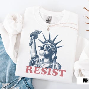 May include: White sweatshirt featuring a graphic of the Statue of Liberty wearing heart-shaped sunglasses and blowing a bubble. The word "RESIST" is printed in red below the image. The sweatshirt is paired with blue jeans.