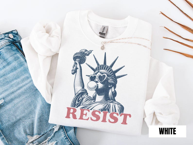 May include: White sweatshirt featuring a graphic of the Statue of Liberty wearing heart-shaped sunglasses and blowing a bubble. The word "RESIST" is printed in red below the image. The sweatshirt is paired with blue jeans.
