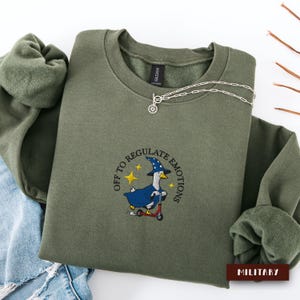 Può includere: Una felpa girocollo verde oliva con un ricamo raffigurante un'anatra bianca che indossa un cappello e un mantello da mago blu mentre guida un monopattino rosso. Il testo "OFF TO REGULATE EMOTIONS" è arcuato sopra con stelle gialle.