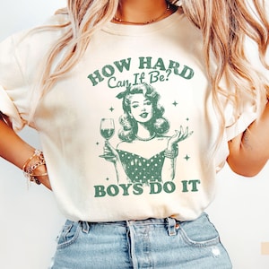 Pode incluir: Camiseta cor marfim com um gráfico verde de uma mulher segurando uma taça de vinho. O texto acima da imagem diz "HOW HARD CAN IT BE?" e abaixo "BOYS DO IT". A mulher está usando um vestido de bolinhas.