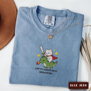May include: A blue jean coloured t-shirt featuring an embroidered graphic of a white cat with a red cape holding a sword while riding a green dragon. The text "OFF TO REINFORCE BEHAVIORS" is embroidered below the graphic in black lettering.