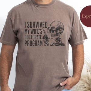 Peut inclure: Un t-shirt marron avec un squelette portant des lunettes de soleil et le texte "J'ai survécu au doctorat de ma femme".