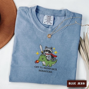 May include: A blue jean coloured t-shirt featuring an embroidered graphic of a raccoon with a sword riding a green dragon. The text below the graphic reads "OFF TO REINFORCE BEHAVIORS". The shirt has a Comfort Colors label inside the collar.