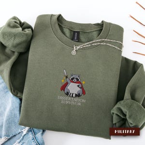 Op de afbeelding: Een olijfgroene sweatshirt met een bruine rechthoek met de tekst "MILITARY". De sweatshirt heeft een geborduurde wasbeer met een cape en een zwaard, met de tekst "DISSERTATION SURVIVOR" eronder. Een zilveren ketting is over de sweatshirt gedrapeerd.