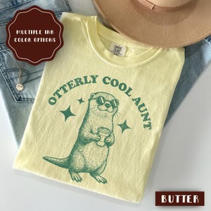 May include: A light yellow t-shirt with the text "Otterly Cool Aunt" and a graphic of an otter wearing sunglasses and holding a coffee cup. The shirt is labeled "Butter" and has multiple ink color options.