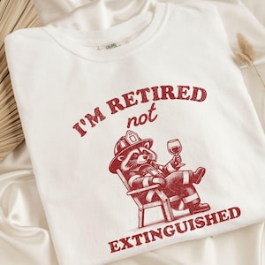 May include: White t-shirt with red text that reads "I'm retired not extinguished". A cartoon raccoon wearing a firefighter's hat and holding a glass of wine is sitting in a chair.