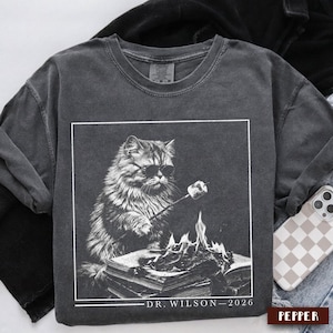 Puede incluir: Sudadera gris oscuro con un gráfico en blanco y negro de un gato con gafas de sol asando una nube de azúcar sobre un libro en llamas. El texto "DR. WILSON - 2026" está impreso debajo. La sudadera tiene un aspecto vintage.