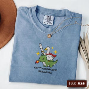 May include: A blue jean coloured t-shirt featuring an embroidered graphic of a white duck wearing a red cape and holding a sword while riding a green dragon. The text "OFF TO REINFORCE BEHAVIORS" is embroidered below the graphic in black lettering.