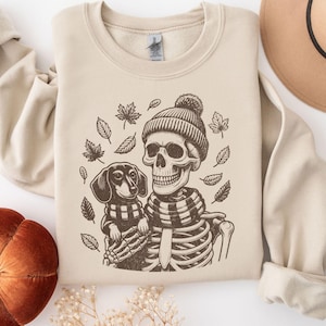 May include: A beige sweatshirt with a graphic print of a skeleton wearing a beanie and scarf holding a dachshund. The skeleton and dog are surrounded by autumn leaves.