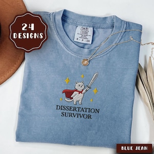 May include: A blue jean coloured t-shirt with an embroidered design. The design features a cat wearing a red cape and holding a sword, with the text "DISSERTATION SURVIVOR" below. A gold necklace is draped across the shirt.