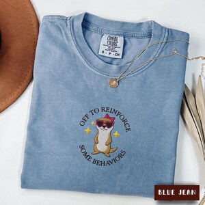 May include: A blue jean coloured t-shirt featuring an embroidered graphic of an otter wearing pink heart-shaped sunglasses and a pink bow. The text reads OFF TO REINFORCE SOME BEHAVIORS with two yellow stars. The shirt has a Comfort Colors brand label.