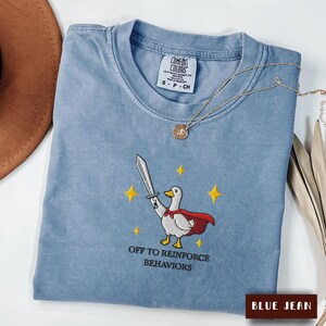May include: A blue jean coloured t-shirt featuring an embroidered graphic of a white goose wearing a red cape and holding a silver sword. The text "OFF TO REINFORCE BEHAVIORS" is embroidered below the goose, surrounded by small yellow stars.