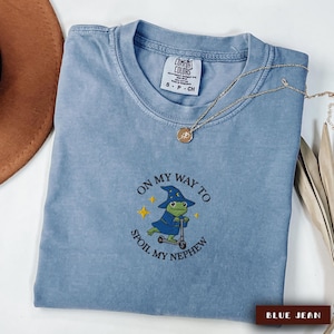 May include: A blue jean Comfort Colors t-shirt with the text "ON MY WAY TO SPOIL MY NEPHEW" and an embroidered frog wearing a wizard hat and riding a scooter. A gold necklace with a pendant is draped across the shirt.