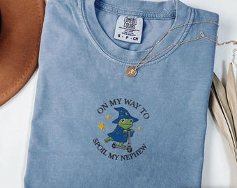 May include: A blue jean Comfort Colors t-shirt with the text "ON MY WAY TO SPOIL MY NEPHEW" and an embroidered frog wearing a wizard hat and riding a scooter. A gold necklace with a pendant is draped across the shirt.