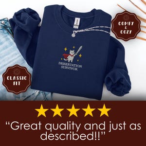 May include: Navy blue sweatshirt with an embroidered design featuring a cat in a cape holding a sword, with the text "Dissertation Survivor." The sweatshirt has a classic fit and is labeled "Comfy & Cozy."