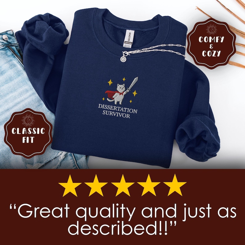 May include: Navy blue sweatshirt with an embroidered design featuring a cat in a cape holding a sword, with the text "Dissertation Survivor." The sweatshirt has a classic fit and is labeled "Comfy & Cozy."