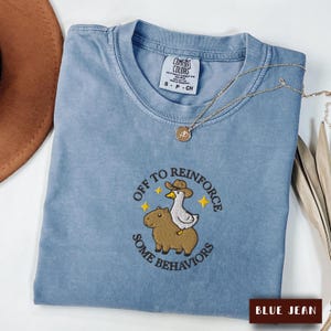 May include: A blue jean coloured t-shirt featuring an embroidered graphic of a capybara with a duck wearing a cowboy hat on its back. The text "OFF TO REINFORCE SOME BEHAVIORS" is arched around the graphic with small yellow stars. A gold necklace rests on the collar.