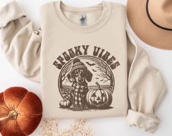 May include: A beige sweatshirt featuring a brown graphic of a dachshund wearing a hat and scarf. The graphic is encircled by the text "Spooky Vibes".