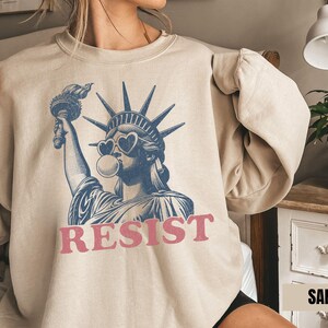 May include: Sand-colored sweatshirt featuring a blue and white graphic of the Statue of Liberty wearing heart-shaped sunglasses and blowing a bubble. The word "RESIST" is printed in pink below the graphic.