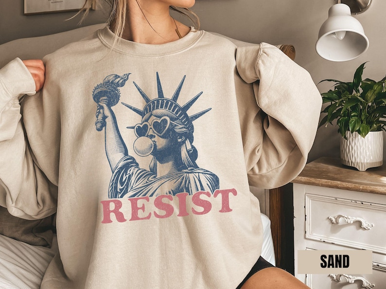 May include: Sand-colored sweatshirt featuring a blue and white graphic of the Statue of Liberty wearing heart-shaped sunglasses and blowing a bubble. The word "RESIST" is printed in pink below the graphic.
