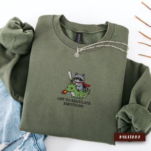 Op de afbeelding: Een olijfgroene sweater met ronde hals en een geborduurde afbeelding van een wasbeer met een rode cape en een zwaard op een groene draak. De tekst onder de afbeelding luidt OFF TO REGULATE EMOTIONS in zwarte letters op de stof.