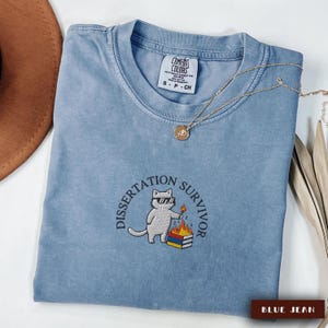 May include: A blue jean Comfort Colors t-shirt with the embroidered text "Dissertation Survivor" and a cartoon cat wearing sunglasses and holding a match. A gold necklace with a pendant is on the shirt.