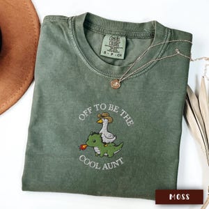 May include: A moss green Comfort Colors t-shirt with the phrase "Off to be the Cool Aunt" and a graphic of a goose wearing a cowboy hat riding a green dinosaur. A gold necklace is draped across the shirt.