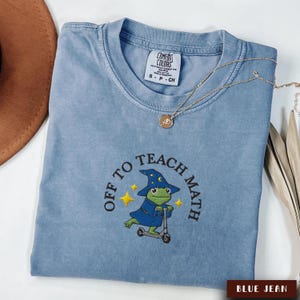 May include: Blue jean Comfort Colors t-shirt with the embroidered phrase "OFF TO TEACH MATH" and a frog wearing a wizard hat and robe riding a scooter. A gold necklace and a brown hat are also visible.
