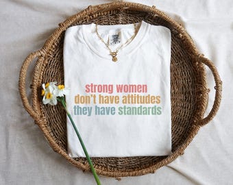 Pode incluir: Uma camiseta branca com o texto "strong women don't have attitudes they have standards" em cores pastel. A camiseta está em uma bandeja tecida com um colar de ouro e uma flor.