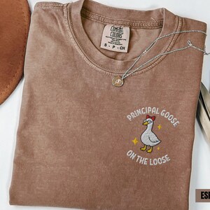 May include: Espresso-colored t-shirt with the embroidered phrase "Principal Goose on the Loose" and a cartoon goose wearing a bow tie. A silver necklace with a pendant is draped across the shirt.