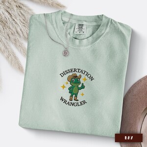 May include: A pale green t-shirt featuring the words "Dissertation Wrangler" and a cartoon frog in a cowboy hat. A silver necklace with a circular pendant is also visible.