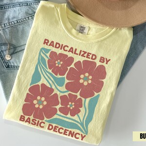 Op de afbeelding: Een lichtgeel t-shirt met de tekst "RADICALIZED BY BASIC DECENCY" in het rood. Het shirt heeft een bloemenontwerp in rood en blauwgroen. Het t-shirt is opgevouwen op een blauwe spijkerbroek en een beige hoed.