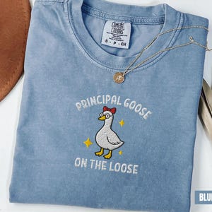 May include: Blue jean-colored t-shirt with an embroidered design. The design features a cartoon goose wearing glasses and a red bow, with the text "PRINCIPAL GOOSE ON THE LOOSE". A gold necklace with a pendant is draped across the shirt.