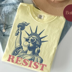 May include: A light yellow t-shirt featuring a graphic of the Statue of Liberty wearing heart-shaped sunglasses and blowing a bubble. The word "RESIST" is printed in red. The shirt is folded on top of blue jeans.