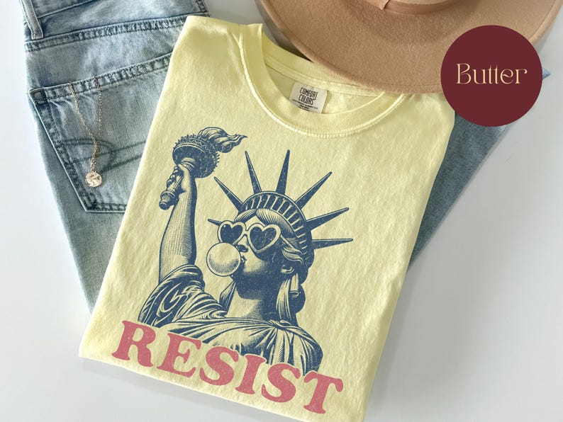 May include: A light yellow t-shirt featuring a graphic of the Statue of Liberty wearing heart-shaped sunglasses and blowing a bubble. The word "RESIST" is printed in red. The shirt is folded on top of blue jeans.