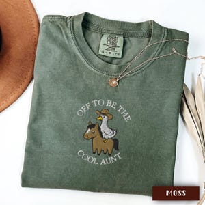May include: A moss green Comfort Colors t-shirt with the embroidered phrase "Off to be the Cool Aunt". The design features a horse and a duck wearing a cowboy hat. A gold necklace is draped across the shirt.