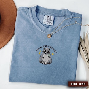 May include: A blue jean Comfort Colors t-shirt with the embroidered phrase "Feral and Engaged" and a raccoon graphic holding a cocktail. A gold necklace with a pendant is draped on the shirt.