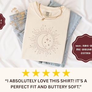 May include: Cream-colored t-shirt with a celestial design featuring a crescent moon, stars, and sunburst. The shirt is folded over blue jeans. The text on the shirt reads "COMFORT COLORS" and "100% RING-SPUN PRE-SHRUNKEN COTTON".