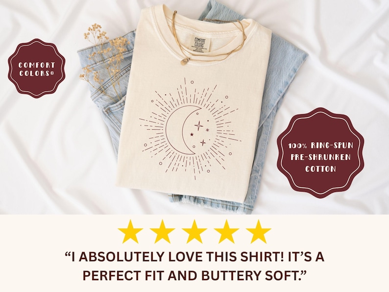 May include: Cream-colored t-shirt with a celestial design featuring a crescent moon, stars, and sunburst. The shirt is folded over blue jeans. The text on the shirt reads "COMFORT COLORS" and "100% RING-SPUN PRE-SHRUNKEN COTTON".